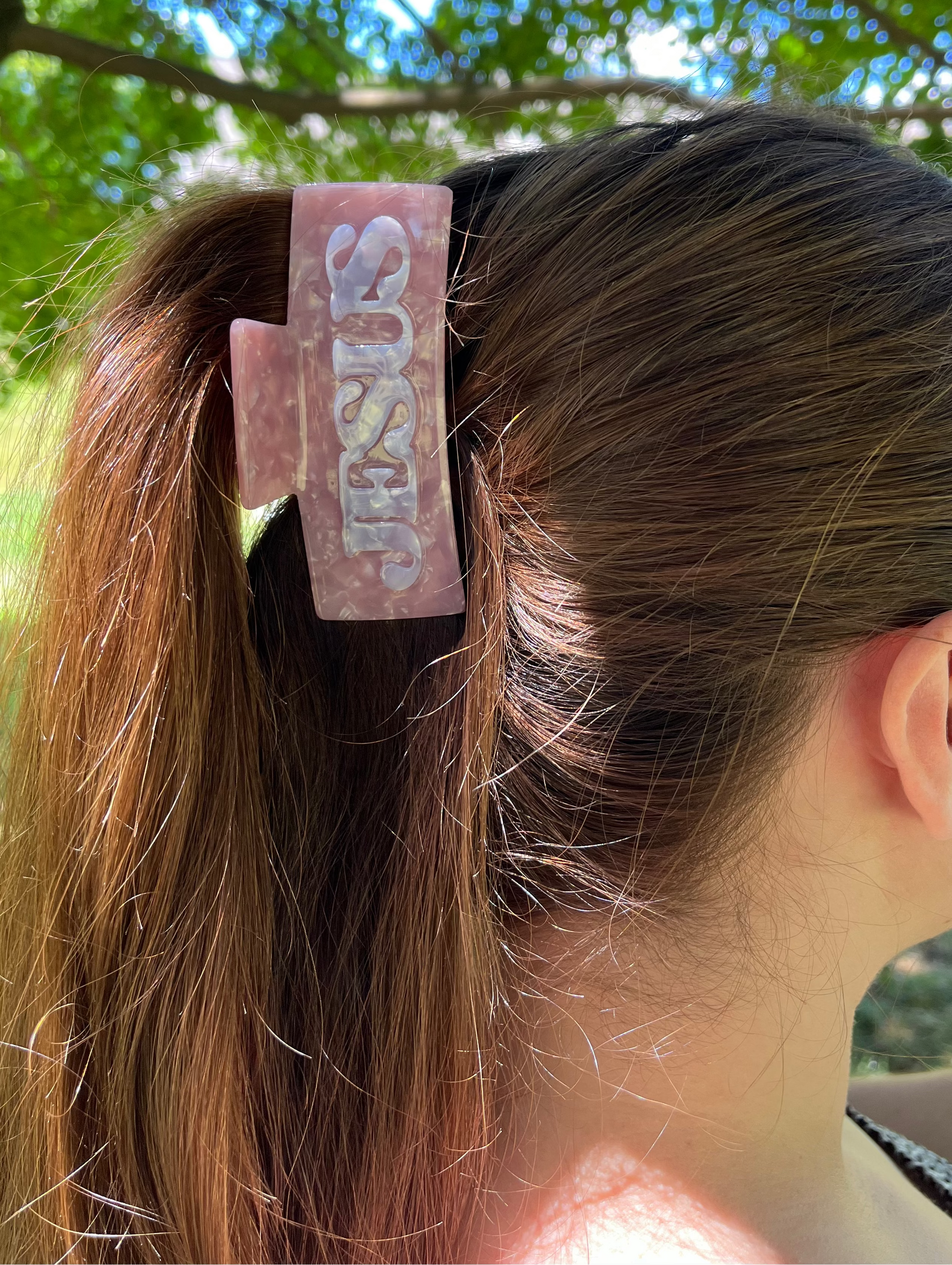 A person outdoors wearing a large rectangular hair claw clip in a dusty pink color with the word 'SUSY' embossed on the front, holding up a brown ponytail.