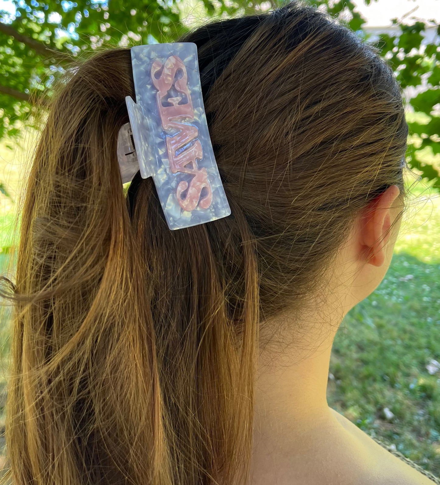 A woman outdoors with her hair in a high ponytail, wearing a decorative translucent hair clip with pink lettering that reads 'SWEETHEARTED'.